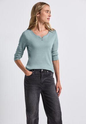 Haut bleu clair à manches longues avec un col henley et deux boutons à l'avant, associé à un jean taille haute gris foncé. Tissu texturé.