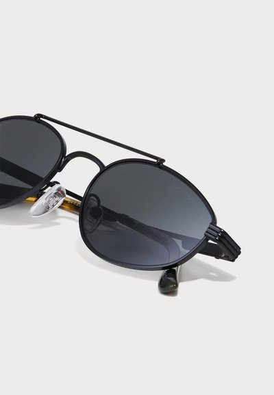Lunettes de soleil à monture noire avec des verres ronds surdimensionnés, un pont en métal et des embouts de branches texturés noirs. Verres teints sombres.