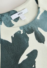 Närbild av en klännings halsringning med blå och vita abstrakta blommönster samt en sydd vit etikett med texten "NEXT."