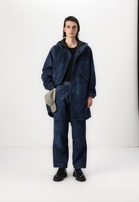 Jaka ar jūras spēku rakstu un kapuci, saskaņota ar atbilstošām biksēm un strukturētu audumu; modelis kreisajā rokā tur pelēku aksesuāru.