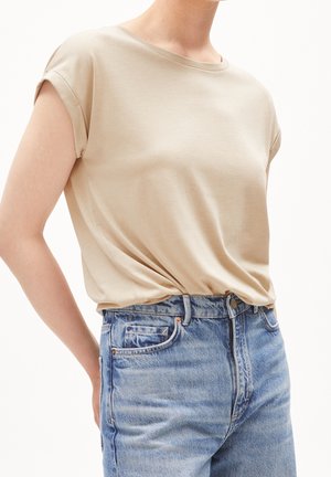 Persona que lleva una camiseta de manga corta color beige, metida ligeramente dentro de unos vaqueros de mezclilla de tiro alto y color azul claro, sobre un fondo blanco.