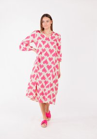 Fließendes beiges Kleid mit pinken Blattmustern, V-Ausschnitt und 3/4-Ärmeln. Kombiniert mit pinken, geknoteten Sandalen für einen abgestimmten Look.