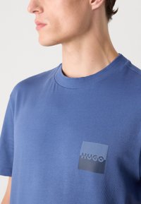 Zila kokvilnas T-kreklā ar apaļu kaklu un īsām piedurknēm. Uz kreisās krūts daļas ir kvadrāta formas logo uzplecs tumšāk zils.