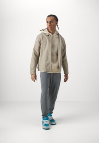 Beige hættejakke over en lys trøje, parret med grå sweatpants og blå sneakers. Jakken har en frontlynlås og lommer.