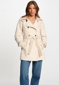 Manteau trench beige ceinturé avec un design croisé, grand col et poches latérales, fabriqué dans un tissu lisse.