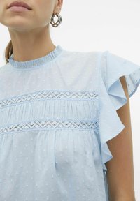 Blusa azzurro chiaro realizzata in tessuto testurizzato con motivi a pois. Presenta maniche arricciate e dettagli in pizzo lungo il petto.