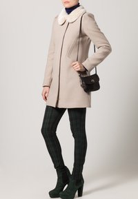 Beige ullkappa med fuskpälskrage, enkelknäppt design, kombinerad med gröna rutiga leggings och gröna platåskor i mocka. Svart axelremsväska.