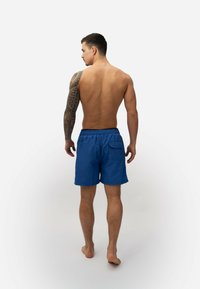 Blauwe zwembroek met een elastische tailleband, achterzak en een gladde textuur, weergegeven vanaf de achterkant op een model met blote huid.