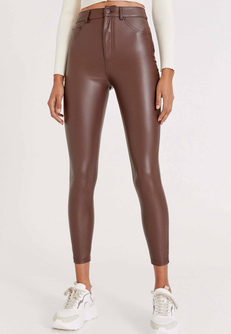 Femme portant des leggings marron en simili cuir taille haute avec fermeture par bouton et zip, associés à des baskets blanches et un pull léger.