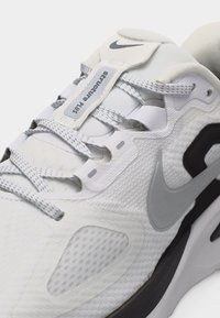 Κοντινό πλάνο λευκού αθλητικού παπουτσιού Nike με γκρι λογότυπο, κορδόνια με μοτίβο και ετικέτα «Struture Plus» στη γλώσσα.