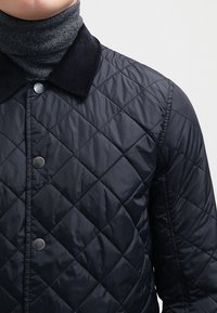 Quiltad svart jacka med rundad manchesterkrage, silverfärgade tryckknappar och en texturerad yta. Grå tröja med hög krage under.