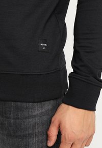 Primo piano del torace di una persona che indossa una felpa nera con un piccolo etichetta "ONLY & SONS" vicino alla vita e jeans grigio scuro.