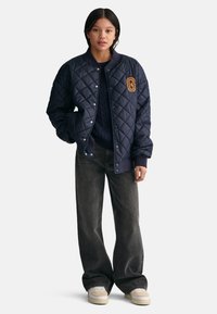 Giubbotto bomber trapuntato blu navy con colletto a costine, che presenta un grande patch arancione a forma di lettera "G". Abbinato a jeans scuri larghi e sneaker color crema.