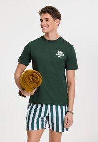 Grünes Baumwoll-T-Shirt mit weißem Text, kombiniert mit grün-weiß gestreiften Badeshorts und einem gerollten Handtuch in Senffarbe.