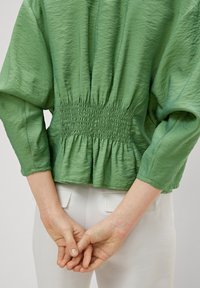 Haut vert texturé avec des manches trois-quarts et un détail froncé à la taille. Porté avec un pantalon blanc, les mains croisées derrière le dos.