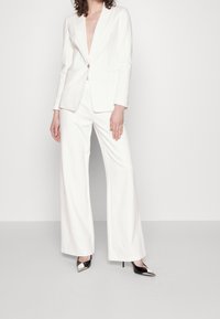 Biały żakiet i szerokie spodnie z gładkiego materiału; żakiet ma głęboki dekolt w kształcie V i jeden złoty guzik, uzupełniony czarnymi i srebrnymi szpilkami.