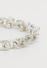 Sudraba ķēdes aproce ar noapaļotiem taisnstūra posmiem un maziem sfēriskiem akcentiem uz balta fona.