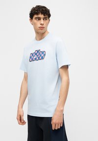 Mladi moški s kratkimi kodrastimi lasmi, ki nosi svetlo modro majico z večbarvnim karo logotipom in temne kratke hlače, gleda levo.