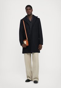 Bărbat îmbrăcat cu un palton negru, cămașă bleumarin, pantaloni bej cu laturi largi, pantofi negri, ținând o geantă crossbody mică în nuanța bej cu curea maro, pe fond alb.