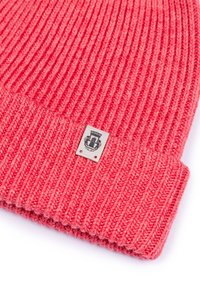 Pink ribbed knit beanie with a folded cuff, featuring a small label with a crest design sewn on the front. Thick, textured yarn.