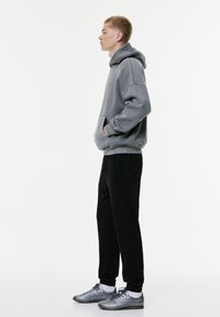 Grijze katoenen hoodie met een kangoeroezak, gecombineerd met zwarte joggingbroek en grijze sportschoenen. Heeft een ontspannen pasvorm en elastische manchetten.