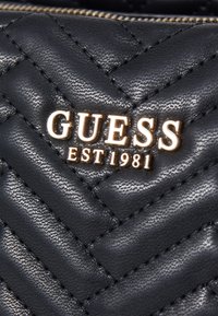 Sort kviltet syntetisk materiale med et chevronmønster og et guld "GUESS EST 1981" logo tydeligt vist.