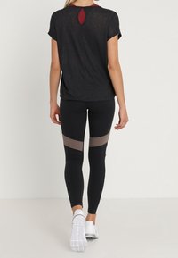 Top atlético negro de manga corta con detalle en forma de llave en la parte trasera, combinado con leggings negros que tienen paneles transparentes en color marrón.
