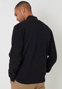 Maglione nero con una texture morbida e colletto alto. Maniche lunghe, polsini e orlo a costine, indossato con pantaloni khaki.