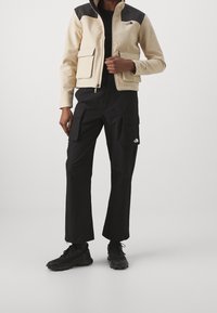 Chaqueta beige con cremallera y paneles de hombros negros, dos bolsillos frontales. Pantalones cargo negros con bolsillos laterales. Zapatos negros con suelas texturizadas.