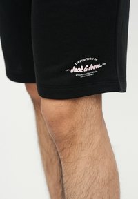 Κοντινό πλάνο ενός τριχωτού ποδιού που φορεί μαύρα σορτς με το λογότυπο και το κείμενο "Jack & Jones" στη ραφή του αριστερού μηρού.