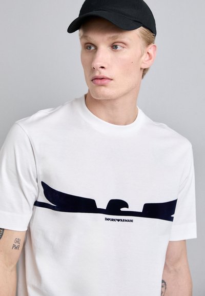 Witte T-shirt van katoen met korte mouwen, voorzien van een donkerblauw embleem en merktekst op de borst. Schoon, eenvoudig ontwerp.