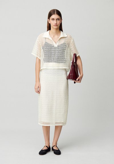 Ensemble haut et jupe en maille à motif quadrillé blanc avec un col. Le mannequin tient un sac à main bordeaux et porte des chaussures plates noires en tissage.