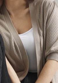 Ljusbeige cardigan med ett strukturerat mönster, öppet fram, och korta ärmar, bärs över en vit linne.