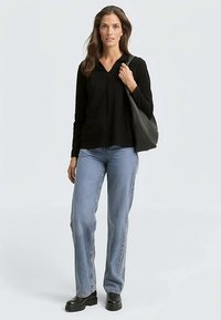 Zwarte top met lange mouwen, een V-hals en subtiele plooidetails, gecombineerd met lichtblauwe jeans en zwarte plateau schoenen, terwijl ze een zwarte tas vasthoudt.