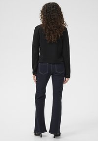 Černá mikina z měkké tkaniny, doplněná tmavě modrými rozšířenými džíny s kontrastním prošitím. Modelka otočená zády, s kudrnatými vlasy.