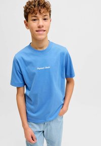 Έφηβος με σγουρά μαλλιά, φορώντας μπλε μπλούζα "Originals Studio" και ανοιχτόχρωμο τζιν, στέκεται με το ένα χέρι στην τσέπη.