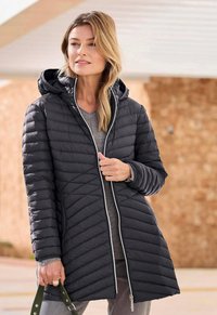 Vrouw in een lange zwarte gewatteerde jas met een capuchon, die een groene tasband vasthoudt, staande in de buitenlucht tegen een vervaagde muur op de achtergrond.