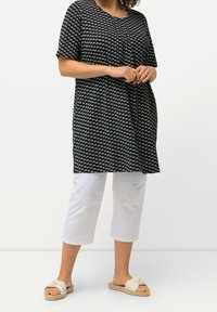 Schwarze Tunika mit weißem geometrischem Muster, kurze Ärmel, runder Ausschnitt. Kombiniert mit weißen, knöchellangen Hosen und hellen Slip-On-Sandalen.
