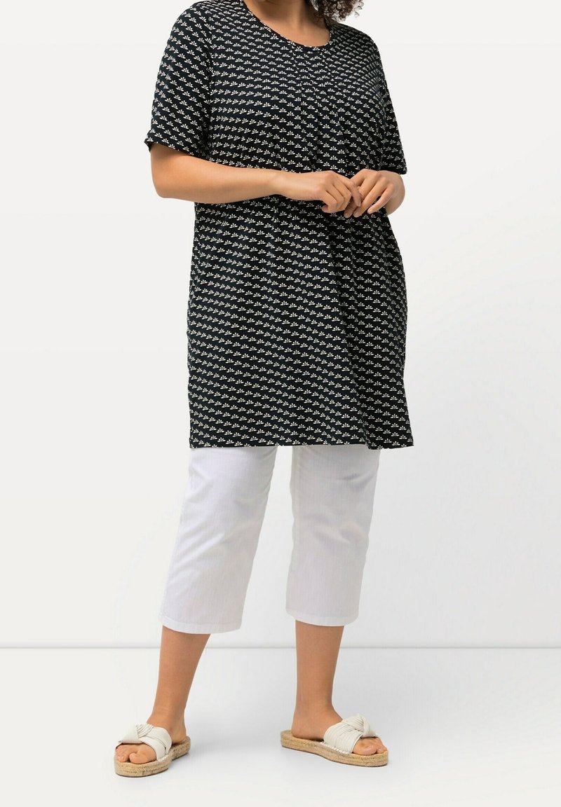 Schwarze Tunika mit weißem geometrischem Muster, kurze Ärmel, runder Ausschnitt. Kombiniert mit weißen, knöchellangen Hosen und hellen Slip-On-Sandalen.