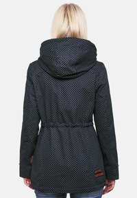 Črno jakno s pikami, ki ima kapuco, zategnjen pas in elastične manšete. Na robu imata dva rjava usnjena detajla. Gladka tekstura materiala.