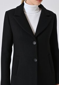 Cappotto di lana nero con colletto a punta, dotato di due tasche anteriori e due bottoni neri. Design minimalista con linee pulite e texture.
