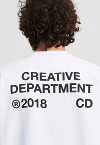 Weißes T-Shirt mit kräftigem schwarzem Text auf der Rückseite, der "CREATIVE DEPARTMENT ® 2018 CD" liest. Weiche Baumwolltextur, lockere Passform.