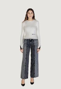 Top blanco de manga larga con textura acanalada y detalles negros, combinado con jeans grises de tiro alto y pierna ancha. Modelo lleva tacones negros, con las manos a los lados.