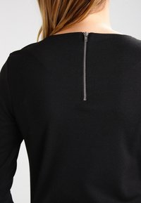 Svart långärmad topp med rak halsringning och silvrig dragkedjestängning i ryggen. Mjukt, slätt tyg.