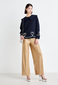 Μπλε πουκάμισο κουμπωτό με ρυτινωτό γιακά και λευκή κεντημένη καρδιά, συνδυασμένο με υφασμάτινο χρυσό παντελόνι σε φαρδιά γραμμή και ασημένια παπούτσια με πλατφόρμα.