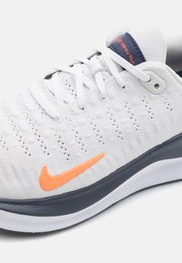 Białe sportowe buty dziane z pomarańczowym znakiem Nike, z teksturowaną cholewką, szarym podszewką i niebieskimi akcentami na języku i wyściółce.