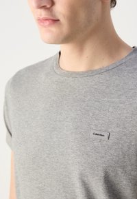 Γκρι βαμβακερό μπλουζάκι με στρογγυλή λαιμόκοψη και κοντά μανίκια. Διαθέτει μια υφαντή ετικέτα Calvin Klein στο στήθος. Απαλή υφή.