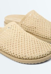 Beige instappersandalen met een geweven bovenzijde en een vlakke, gevoerde zool, getoond vanuit een close-up schuin perspectief.