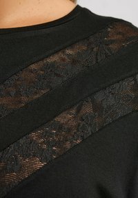 Närbild av en svart tygblus med diagonala genomskinliga spetspaneler med blommönster på axeln och övre bröstet som visar huden under.