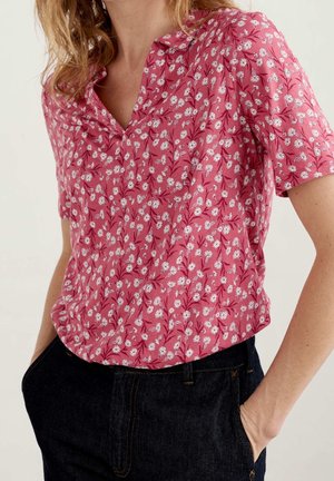 Kvinde iført en lyserød bluse med korte ærmer og hvidt blomsterprint, stukket ned i mørkeblå jeans, hænder i lommerne, foran ensfarvet baggrund.